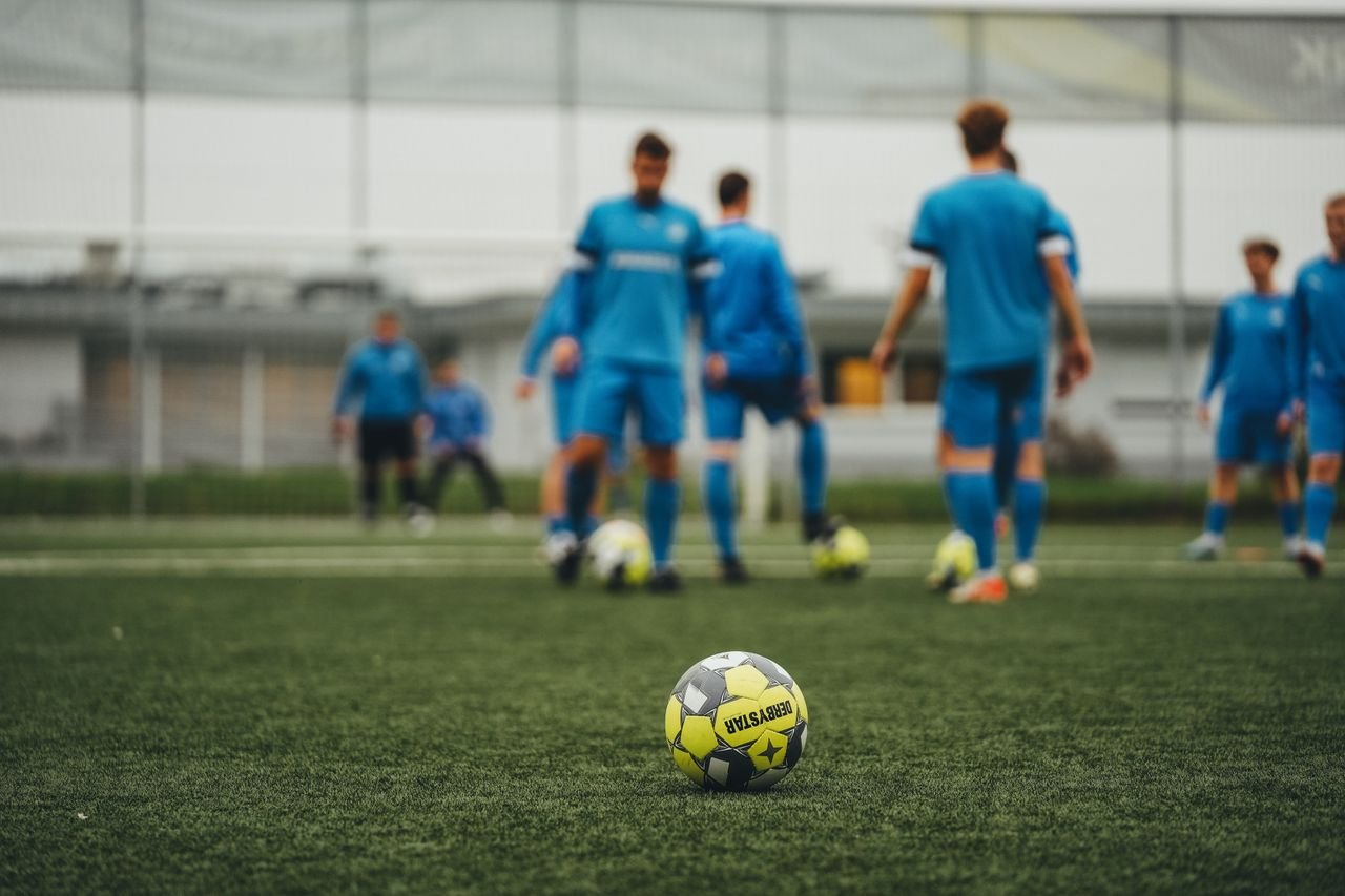 Soccer players in blue uniforms practicing on a green field focus on soccer ball in foreground.